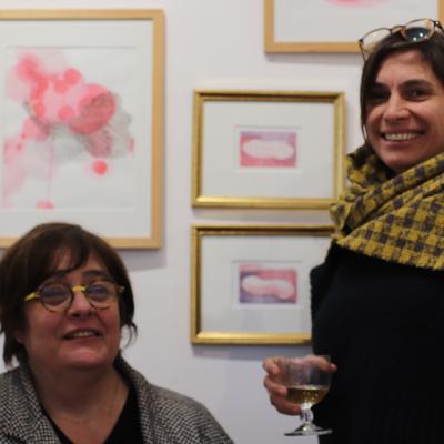 portraits de deux artistes, l'une assise souriant et l'autre tenant un verre souriant également sur fond d'oeuvres aux figures de couleur rose  encadrées