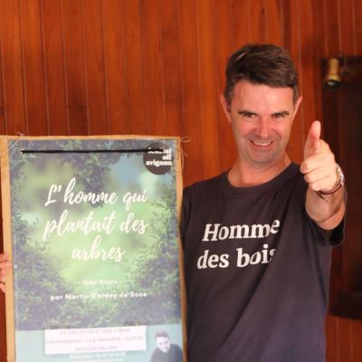 un homme avec un Tshirt où est écrit Homme des bois le pouce levé et un grand sourire. Il tient l'affiche du spectacle