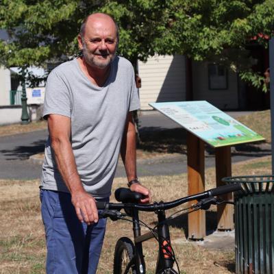 Portrait d'un homme avec son vélo sur fond d'herbe grillée par le soleil et de bâtiments et autres aménagements urbains
