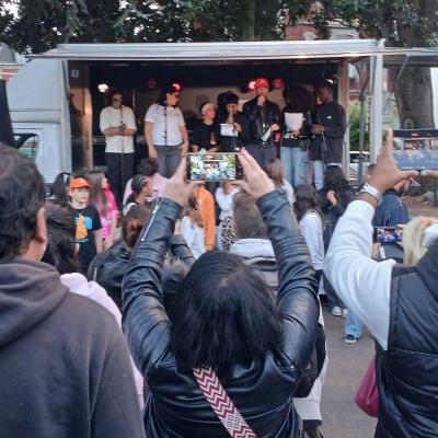 Un camion aménagé en salle de concert avec des jeunes chantant dedans et des personnes de dos prenant des photos du concert avec leur smartphone