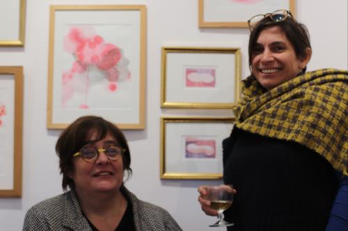 portraits de deux artistes, l'une assise souriant et l'autre tenant un verre souriant également sur fond d'oeuvres aux figures de couleur rose  encadrées