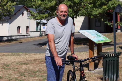 Portrait d'un homme avec son vélo sur fond d'herbe grillée par le soleil et de bâtiments et autres aménagements urbains