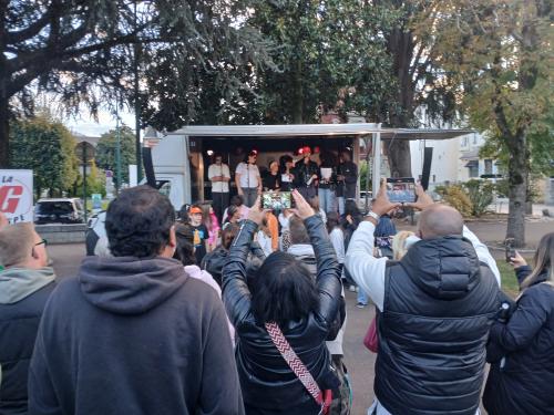 Un camion aménagé en salle de concert avec des jeunes chantant dedans et des personnes de dos prenant des photos du concert avec leur smartphone