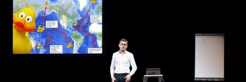 un homme en chemise blanche devant un slide de powerpoint avec une carte du monde et un gros canard jaune