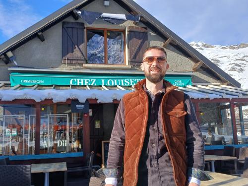 le propriétaire avec ses lunettes de soleil pose devant la devanture de son restaurant entouré de montagnes sous un ciel bleu